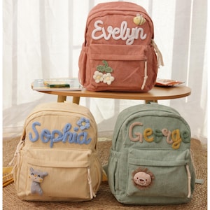 Może przedstawiać: Trzy sztruksowe plecaki w kolorze brzoskwiniowym, żółtym i zielonym. Każdy plecak ma imię w kontrastowym kolorze: "Evelyn" z pszczołą, "Sophia" z kwiatem i "George" z lwem. Posiadają przednie kieszenie i regulowane paski.