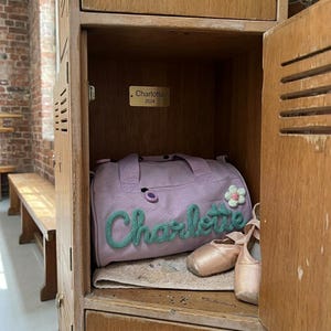May include: A lilac duffel bag with the name "Charlotte" in green yarn, a white and green flower, and ballet shoes inside a wooden locker. A small gold nameplate reads "Charlotte 2024".
