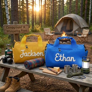 May include: A campsite scene featuring a yellow and a blue duffel bag, each with a name embroidered on it. A rolled plaid blanket, a cutting board with cheese and a knife, and camping gear are on a wooden picnic table. A tent and campfire are in the background.