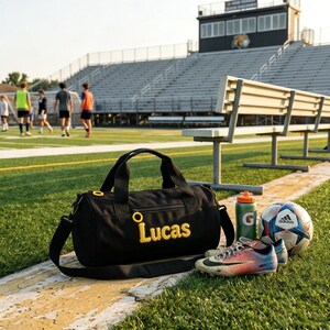 May include: Black duffel bag embroidered with the name "Lucas" in yellow, a football, a pair of football boots, and a water bottle on a grassy pitch. The background shows a stadium with seating and people playing football.