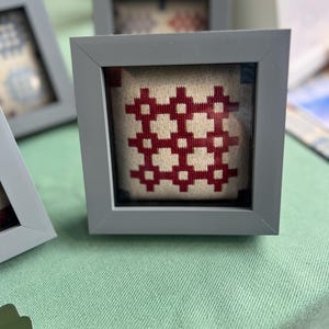 May include: Small gray framed artwork with a red and cream-colored geometric pattern. The artwork is displayed on a light green surface. The frame is square and the artwork is centered within the frame.