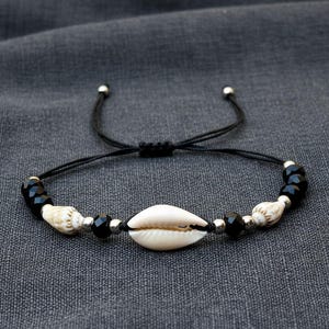 Pode incluir: Pulseira ajustável de cordão preto com uma concha branca central, ladeada por contas pretas e conchas menores. Contas de metal prateado acentuam o design. A pulseira está sobre um fundo cinza.
