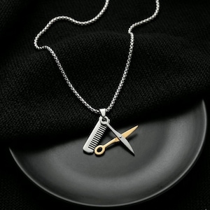 May include: Silver necklace with a pendant depicting a comb and scissors. The comb is silver, and the scissors are gold and silver. The necklace is presented on a black surface.