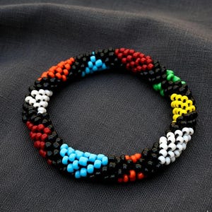 Könnte beinhalten: Ein Armband aus Perlen mit schwarzer Basis und Abschnitten aus roten, orangen, blauen, grünen, gelben und weißen Perlen. Das Armband ist kreisförmig und liegt auf einem dunkelgrauen Stoff. Die Perlen sind klein und eng gewebt.
