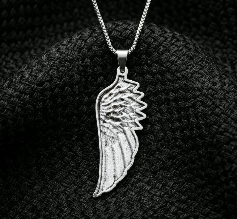 May include: A silver-toned metal pendant shaped like a single bird wing, featuring detailed feather textures and a smooth border. The pendant hangs from a matching silver-toned box chain necklace, showcasing a metallic finish and a structured design.