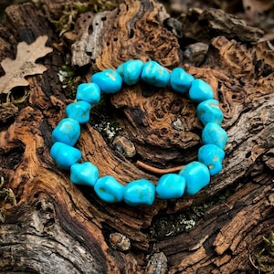 May include: A bracelet made of turquoise beads. The light blue beads are rounded and show natural colour variations. The bracelet is strung together, forming a circle. It sits on a textured, brown wooden surface.