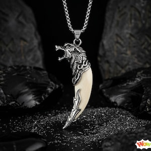 May include: A silver-colored wolf head pendant with an open mouth, attached to a cream-colored tooth-shaped charm. The pendant hangs from a silver chain. The background is dark with black rock formations.