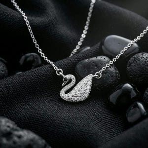 May include: A silver-toned necklace featuring a swan-shaped pendant. The swan body is encrusted with numerous small, clear, sparkling stones, while the eye is accented with a single, small, dark stone. The pendant hangs from a delicate silver chain.