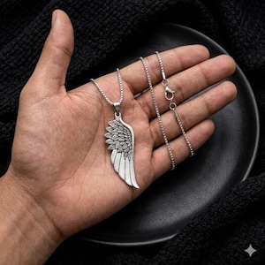 May include: A silver-toned metal pendant shaped like a single angel wing with detailed feather engravings, suspended from a thin silver-toned box chain necklace. The pendant features a polished finish with darkened recessed areas to highlight the texture.