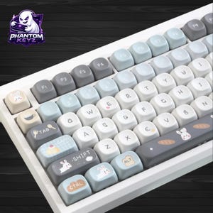 Può includere: Una tastiera meccanica bianca con tasti personalizzati. I tasti sono grigi, azzurri e bianchi, con icone e illustrazioni, tra cui un coniglio e una tazza di caffè. Il logo "Phantom Keyz" è visibile in alto a sinistra.