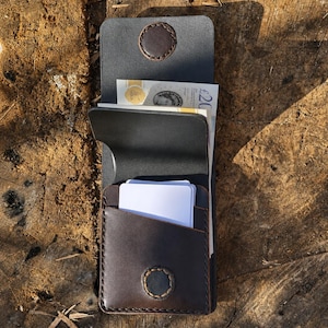 Puede incluir: Una cartera de cuero marrón oscuro con una ranura vertical para tarjetas blancas y un compartimento principal que contiene un billete de veinte libras. La cartera tiene una solapa asegurada con un cierre de botón a presión cosido.