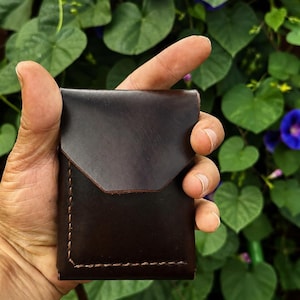 Puede incluir: Un tarjetero de cuero marrón oscuro con un cierre de solapa puntiagudo y costuras gruesas visibles en color marrón a lo largo de los bordes inferiores y laterales. El cuero tiene un acabado liso y mate con forma rectangular.