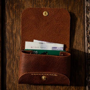 May include: A brown leather wallet with a flap closure secured by a brass snap button. The wallet contains a green card and a white paper bill with the visible text CH83 895178. The leather features visible stitching along the bottom edge.
