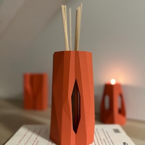 May include: An orange geometric diffuser with reeds, a candle holder, and a small container are on a book. The diffuser has a diamond-shaped opening. The candle holder has a cutout design. The items are on a book.