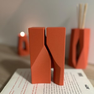 Könnte beinhalten: Eine orangefarbene geometrische Skulptur mit geteiltem Design, die auf einem Buch mit Text steht. Im Hintergrund sind eine Kerze und ein Schilfrohrdiffusor zu sehen. Die Objekte sind modern und haben eine minimalistische Ästhetik.