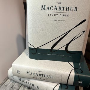 Op de afbeelding: Drie exemplaren van "The MacArthur Study Bible" zijn op elkaar gestapeld. De boeken hebben een lichtgekleurde kaft met de titel in zwart en de woorden "Study Bible" in een kleinere letter. De rug is donkerblauwgroen. Het bovenste boek toont de tekst "Unleashing God's Truth One Verse at a Time."