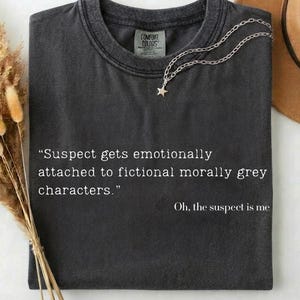 Puede incluir: Camiseta gris oscuro con el texto "Suspect gets emotionally attached to fictional morally grey characters." y "Oh, the suspect is me." Un collar plateado con un colgante de estrella descansa sobre la camiseta.