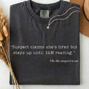 May include: Dark gray t-shirt with the text "Suspect claims she's tired but stays up until 3AM reading. Oh, the suspect is me." A silver necklace with a star pendant rests on the shirt. A brown hat and dried flowers are in the background.