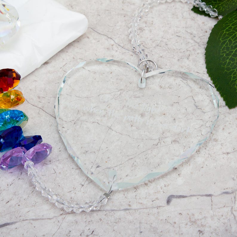May include: A clear, heart-shaped glass ornament with a beaded string for hanging. The ornament has etched text and is surrounded by colorful crystals. A white package and a green leaf are in the background.