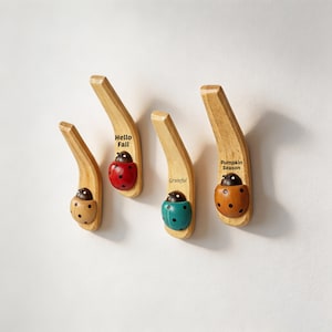 Puede incluir: Cuatro ganchos de pared de madera clara, cada uno con una pequeña figura de mariquita en la base. Las mariquitas son de color beige, rojo, turquesa y naranja. La mariquita roja tiene el texto Hello Fall, la turquesa dice Grateful y la naranja Pumpkin Season.