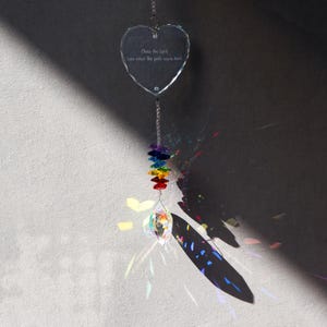 May include: A clear, heart-shaped crystal suncatcher with the text "Chase the light even when the path seems dark." A rainbow of colored crystals hangs below, with a larger crystal at the bottom. The suncatcher is suspended by a silver chain.