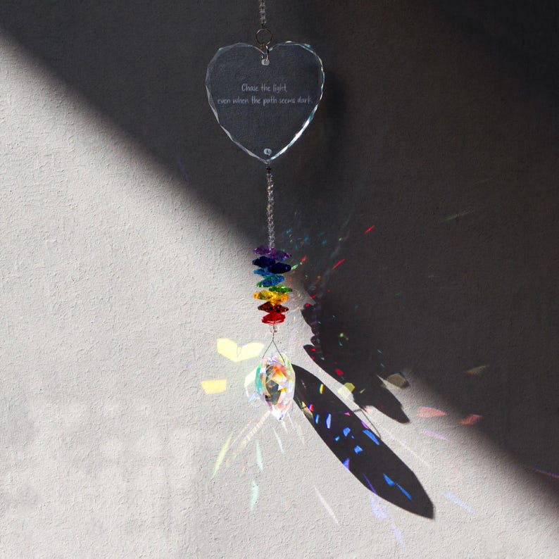 May include: A clear, heart-shaped crystal suncatcher with the text "Chase the light even when the path seems dark." A rainbow of colored crystals hangs below, with a larger crystal at the bottom. The suncatcher is suspended by a silver chain.