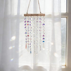 Puede incluir: Un atrapasoles decorativo de cristal colgante con el texto "Love & Light" en una barra de madera. El atrapasoles presenta múltiples hebras de cristales iridiscentes y colgantes transparentes en forma de lágrima, suspendidos por un cordel natural.