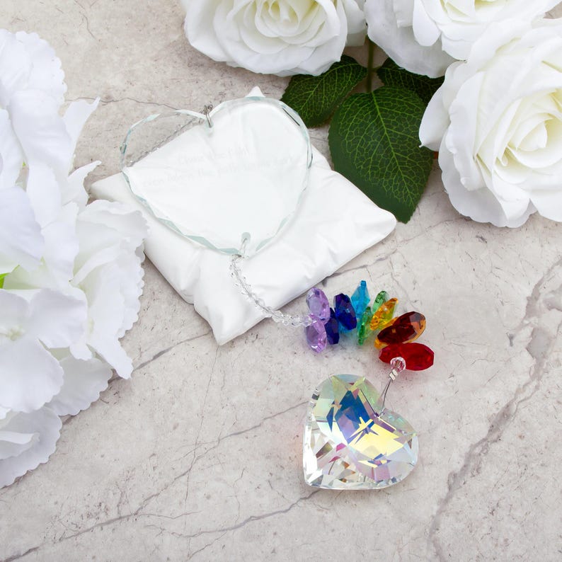 May include: A clear, heart-shaped crystal ornament with a rainbow of colored crystals attached. The ornament is on a white pillow, with white roses in the background. The crystal heart is approximately 3 inches wide.