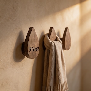 Puede incluir: Tres ganchos de pared de madera con forma de gota alineados. El gancho de la izquierda tiene la palabra Koa grabada en una fuente cursiva oscura. Una bufanda de punto beige con flecos cuelga del gancho central, ocultando su superficie.