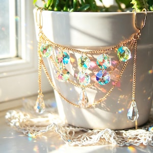 May include: A gold-tone chain with iridescent crystal embellishments hangs in front of a white ceramic pot. The crystals are multi-faceted and reflect light, creating a rainbow effect. The chain is layered, with teardrop crystals suspended below.