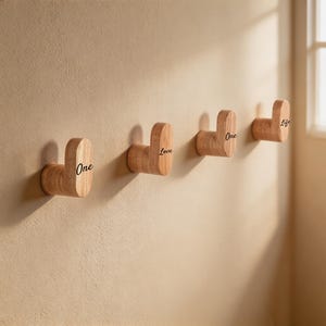 以下が含まれることがあります： 水平に並べて取り付けられた4つの明るい色の木製ウォールフック。各フックは丸みを帯びたL字型のデザインです。フックの側面には黒い文字で「One」「Love」「One」「Life」という言葉がそれぞれプリントされています。