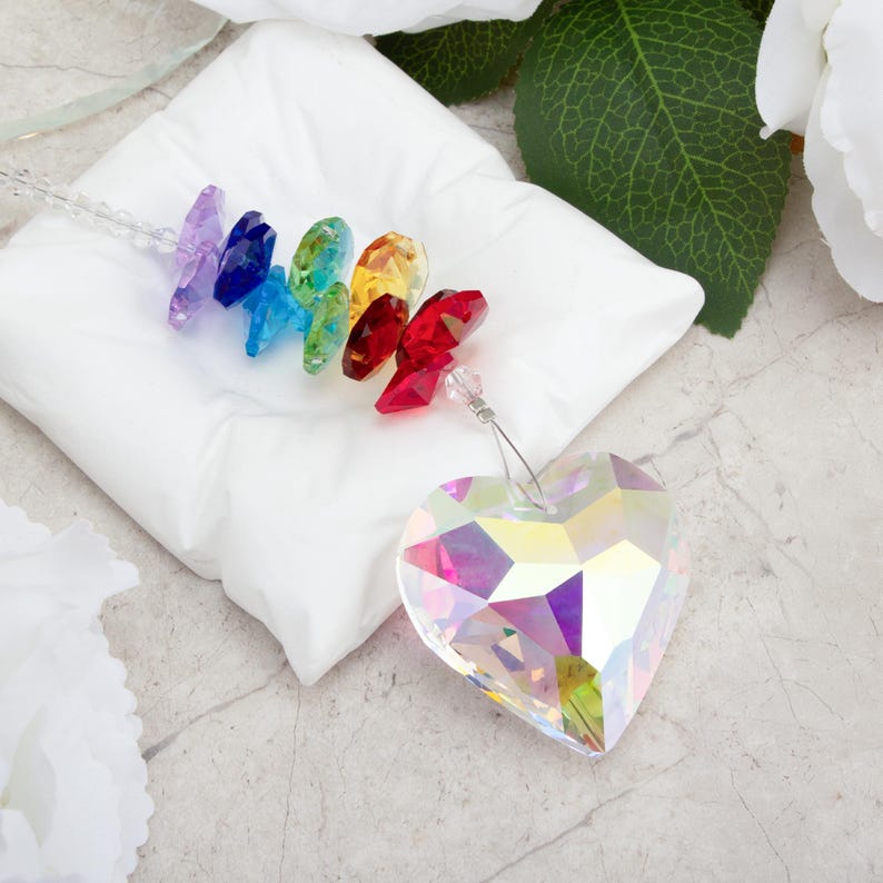 May include: A heart-shaped crystal prism and a rainbow of colorful crystals on a white pillow. The prism reflects multiple colors. The crystals are arranged in a row, showcasing a spectrum of hues.