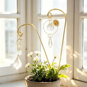 Könnte beinhalten: Ein goldfarbener Pflanzenstecker aus Metall mit himmlischen Kristall- und Metallanhängern. Die Anhänger umfassen eine Sonne, einen Mond und Kristallanhänger. Der Stecker befindet sich in einem Topf mit weißen Blumen und grünem Laub.