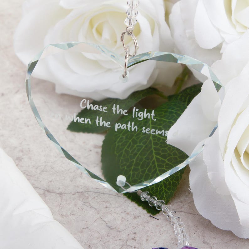 May include: A clear, heart-shaped crystal ornament with the inscription "Chase the light, when the path seems..." is suspended against a backdrop of white roses and green leaves. The ornament hangs from a silver-colored chain.