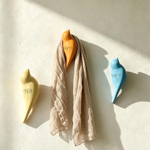 May include: Three bird-shaped wall hooks in yellow, orange, and blue. The yellow hook is labelled 'His', while the orange and blue hooks are labelled 'Hers'. A beige scarf is draped over the orange bird hook, which is mounted on a textured wall.
