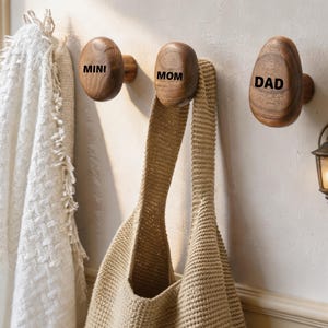 Peut inclure: Trois patères murales en bois naturel de forme arrondie, montées horizontalement. Chaque patère présente une finition lisse et polie avec des motifs de grain de bois foncé. Elles sont gravées avec le texte noir MINI, MOM et DAD, servant d'organisateurs muraux fonctionnels.