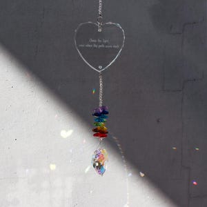 May include: A clear, heart-shaped crystal suncatcher with the text "Chase the light even when the path seems dark." It features a rainbow-colored bead strand and a teardrop-shaped crystal at the bottom, hanging from a silver chain.