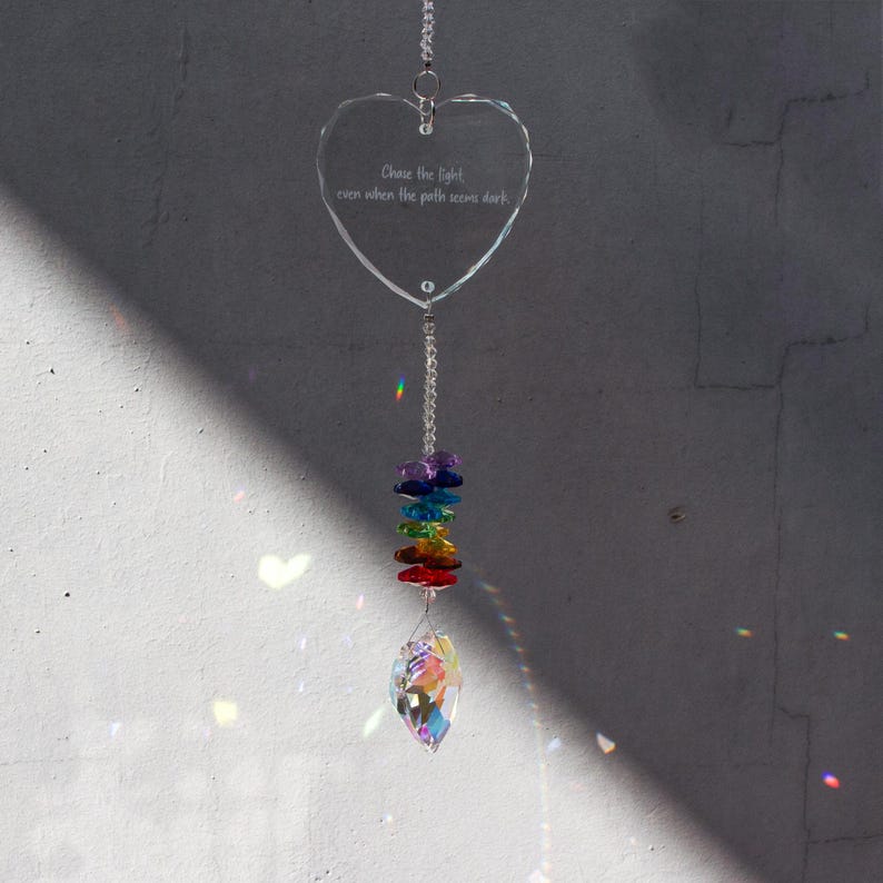May include: A clear, heart-shaped crystal suncatcher with the text "Chase the light even when the path seems dark." It features a rainbow-colored bead strand and a teardrop-shaped crystal at the bottom, hanging from a silver chain.