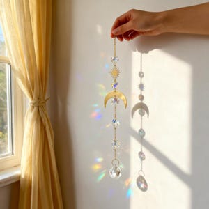 May include: A celestial-themed suncatcher with a gold-colored crescent moon, sun, and crystal accents. The suncatcher is suspended by a delicate chain and is held by a hand. The crystals create a rainbow effect when light shines through them.