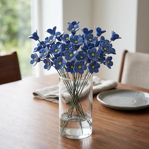 May include: A clear glass vase holds a bouquet of blue ceramic flowers with yellow centers. The flowers are arranged on thin green stems. The vase sits on a wooden table, with a neutral-colored napkin and plate in the background.
