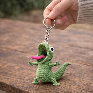 Puede incluir: Un llavero de lagarto verde tejido a ganchillo con una lengua rosa y la boca abierta. El lagarto tiene ojos negros y blancos y está unido a un llavero y una cadena plateados. El llavero se muestra sobre una superficie de madera.