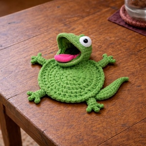 Op de afbeelding: Een handgemaakte groene gehaakte kikker onderzetter met een wijd open mond en een roze tong. De kikker heeft witte en zwarte ogen, kleine armen en benen en een staart. De onderzetter ligt op een houten tafel.