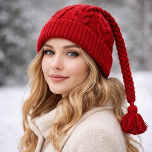 Puede incluir: Un gorro de punto rojo brillante con una larga borla trenzada. El gorro presenta un diseño de punto de cable y un borde acanalado. La mujer de la imagen lleva un abrigo beige y tiene el pelo rubio. El fondo es una escena invernal borrosa.