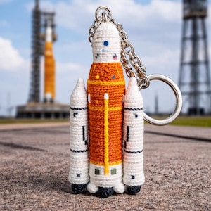 May include: A crocheted rocket ship keyring. The rocket has an orange body, white boosters, and a white top. It's attached to a silver chain and ring. The background shows a launch pad and a rocket.