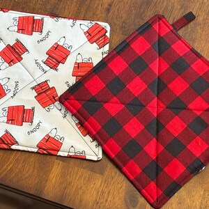 May include: Two square pot holders. One is white with a repeating pattern of Snoopy in his doghouse, with the word "SNOOPY". The other is red and black buffalo plaid.