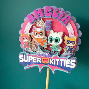 May include: A vibrant cake topper with the name "AMELIA" and the number "5" in glitter. It showcases four cartoon cat characters from Disney Junior's "Super Kitties", with the show's logo at the base, all on a wooden stick.