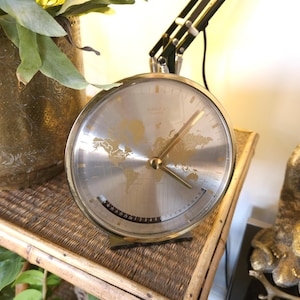 May include: A round analogue desk clock with a metallic silver face featuring a gold-toned world map graphic. The clock has gold-coloured hour, minute, and second hands, and a curved date display window at the bottom with black numbers on a white background.