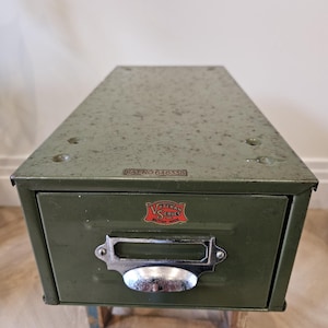 May include: An olive green metal drawer cabinet with a single drawer and a silver handle. The cabinet features the text "PAT NO 646338" and a "Veteran Series British Made" emblem. It rests on a small, multi-coloured wooden stand.