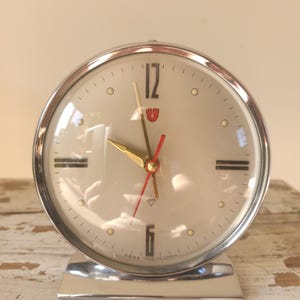 May include: A round analogue table clock with a polished silver-tone metal frame and base. The off-white face features black numerals at 12 and 6, gold-tone hour and minute hands, a red second hand, and a small diamond icon above the 6.
