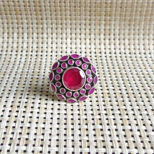 Può includere: Un anello circolare in argento con una grande pietra centrale sfaccettata di colore rosa intenso, circondata da un gruppo di pietre rosa più piccole, rotonde e sfaccettate, incastonate in una montatura in metallo scuro ossidato che contrasta.